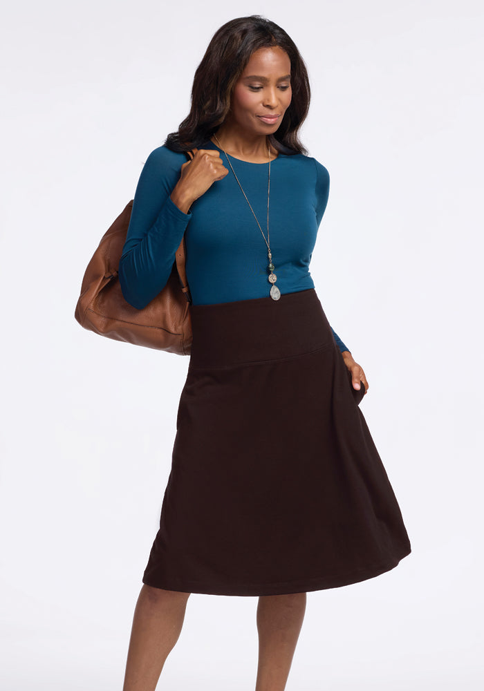 A woman wearing a Woolx blue long-sleeve top and the Marilyn Flowy Midi Skirt in French Roast is holding a brown bag. She accessorizes with a long necklace and stands in a neutral pose against a light background.