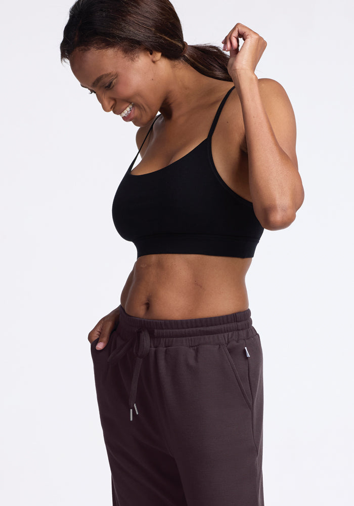 A woman smiles and looks down, wearing a black sports bra paired with the Woolx Parker Sweatpants in Chocolate Plum. In a relaxed pose, she touches her hair against a plain background, enjoying the comfort and temperature-regulating properties that keep her feeling fresh.