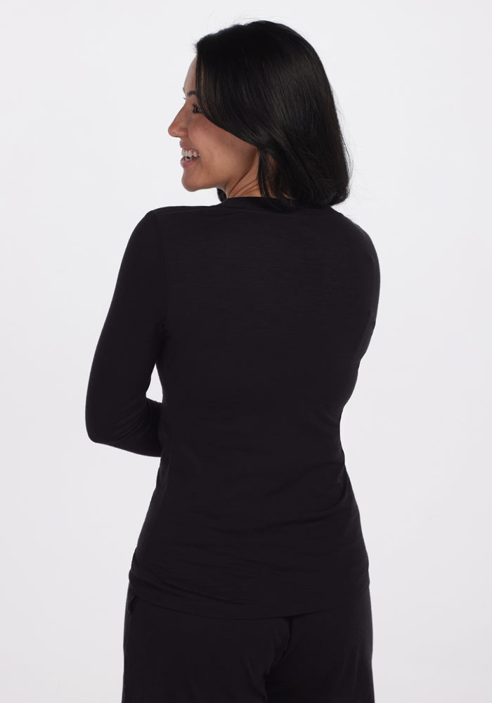 A person with long dark hair is standing and smiling over their shoulder, wearing a comfortable Lily V Neck Lounge Top in black by Woolx and black pants against a plain white background.