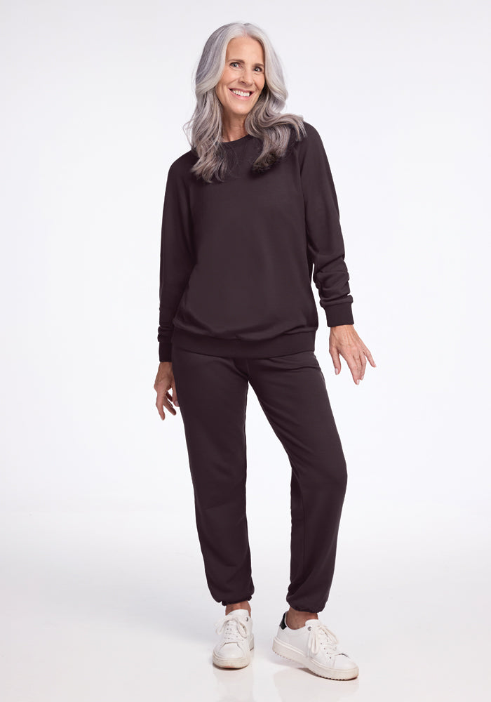 A woman with long gray hair is smiling and posing in the Bailey Crewneck Sweatshirt in Chocolate Plum from Woolx, paired with matching pants and white sneakers. She stands gracefully against a plain white background.