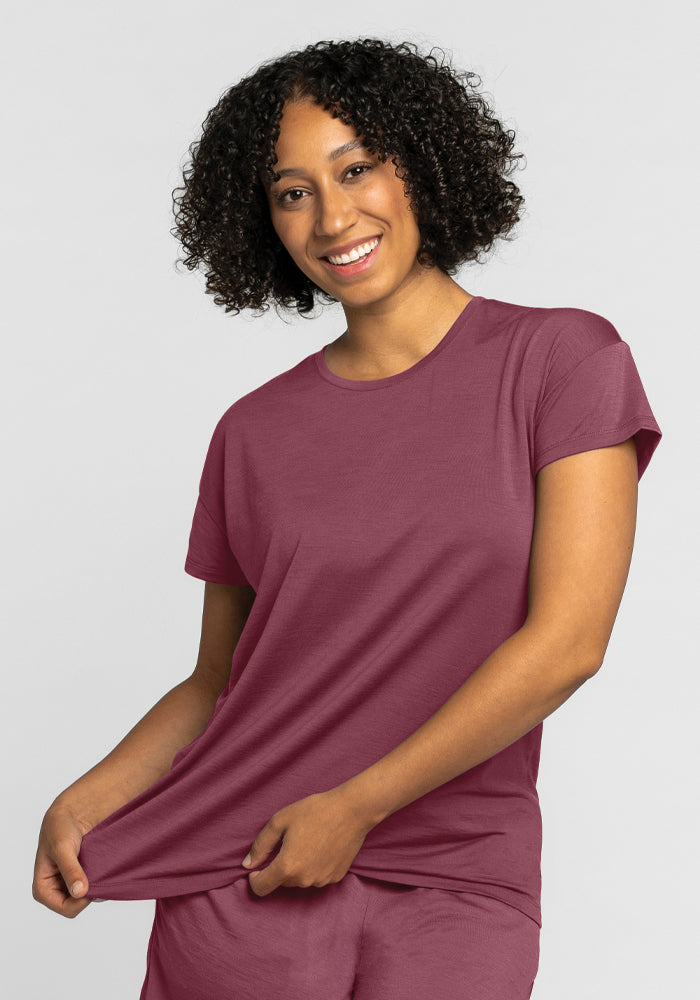 A person with curly hair smiles while wearing a Billie Lounge Tee in a wild ginger hue and matching pants by Woolx. They gently hold the bottom of their shirt, pulling it slightly to the side. The background is plain and neutral.