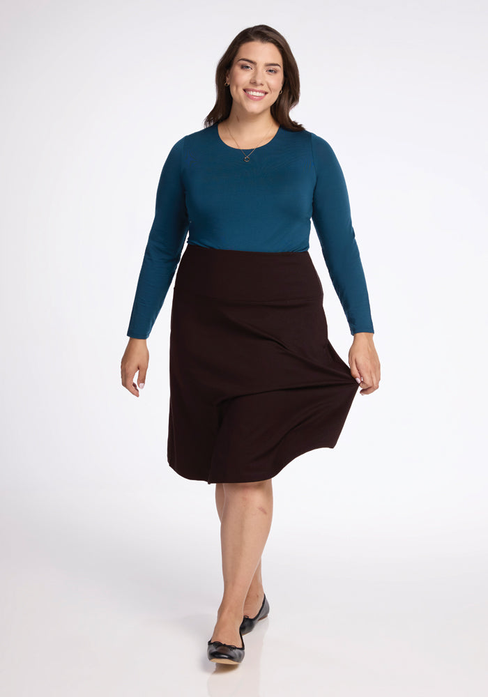 A person dressed in a blue long-sleeve top and the Marilyn Flowy Midi Skirt in French Roast from Woolx is smiling as they walk forward. They have long hair and are sporting black shoes against a plain, light background.