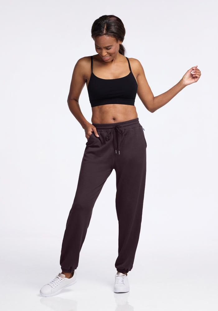 A woman is wearing a black sports bra and Woolx's Parker Sweatpants in Chocolate Plum as she stands against a plain white background. Smiling and looking down, she has one hand raised slightly. Her outfit, paired with white sneakers, offers an odor-free experience while keeping her comfortable throughout the day.