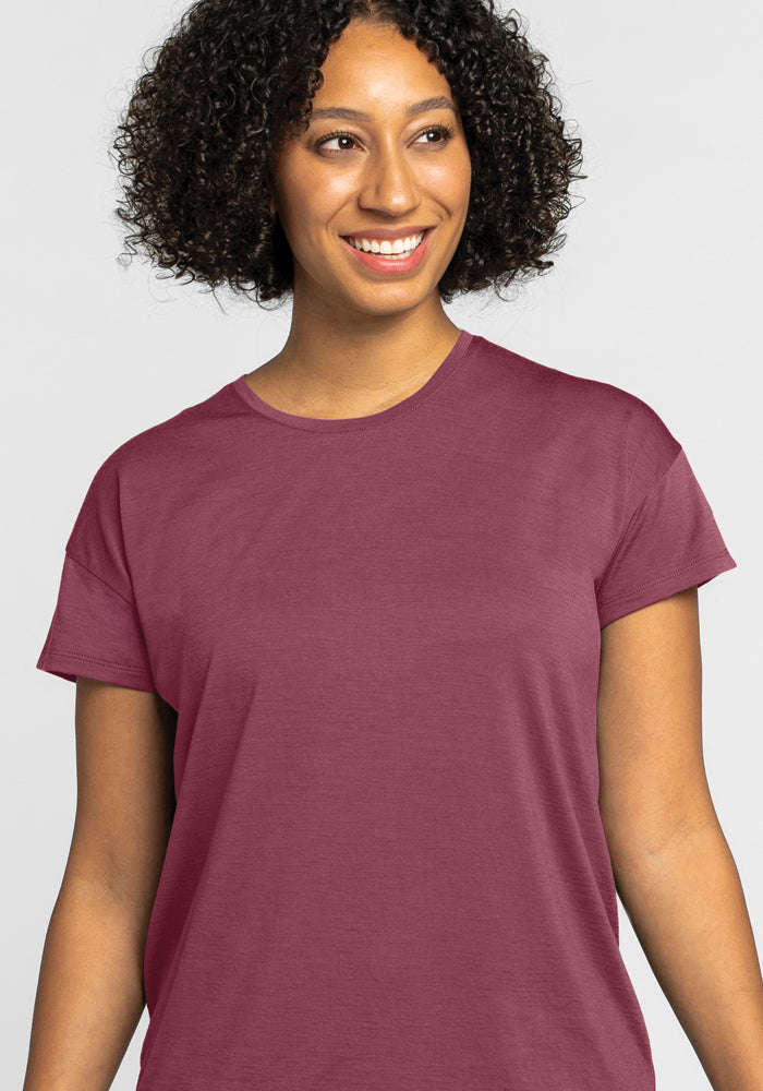 A person with curly hair smiles while wearing the Billie Lounge Tee in Wild Ginger, a temperature-regulating garment by Woolx. The light gray background adds a touch of simplicity.