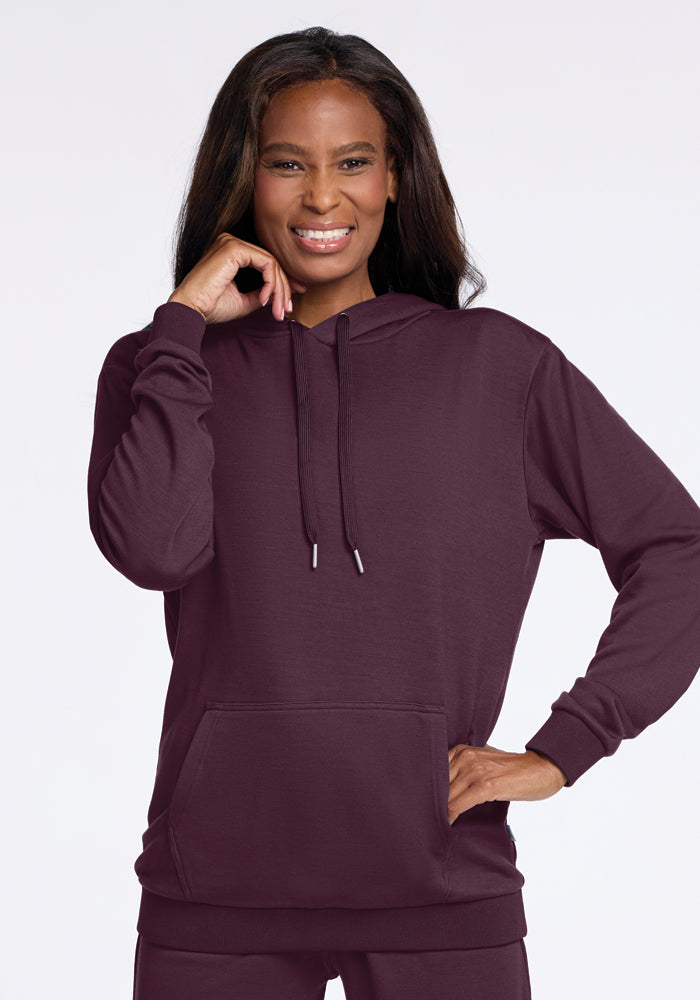 A person smiles while wearing a Wine Tasting Avery Hoodie from Woolx, which features a front pocket. Crafted from soft Merino wool, the hoodie includes drawstrings, and the individual has long, dark hair. They stand against a plain, light-colored background. 