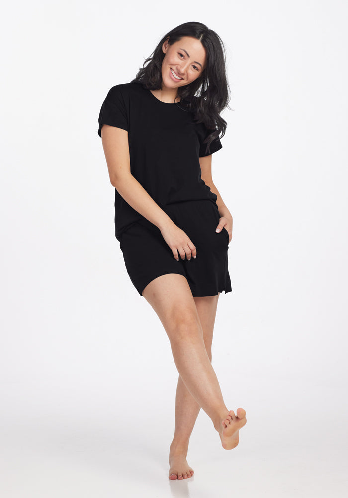 A person with long dark hair is smiling and standing on one leg in a playful pose, wearing the Billie Lounge Tee in black from Woolx, made of moisture-wicking fabric and shorts against a plain white background.