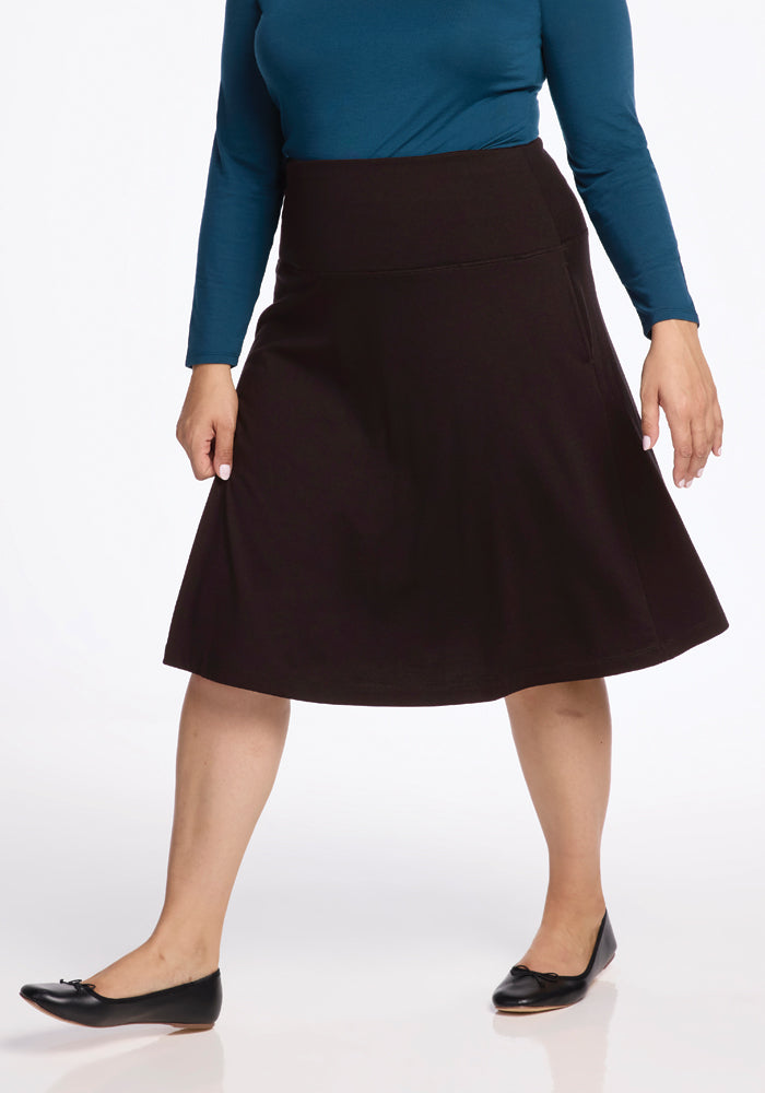 A person wearing a teal long-sleeve top and the Woolx Marilyn Flowy Midi Skirt in French Roast, paired with black flat shoes, walks against a plain white background.