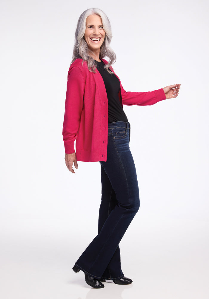 A person with long gray hair is smiling and posing playfully, wearing the Woolx Gia Sweater in Berry Burst, along with a black top, dark jeans, and black shoes against a plain white background.