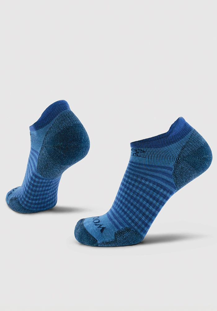 Two Woolx Amelia No Show Tab Lightweight Socks in Blue Lagoon are shown as if being worn, highlighting their cushioned support and textured patterns. The design features raised heels and darker blue accents on the toes, heels, and cuffs against a lighter blue patterned backdrop. They are displayed against a plain white background.