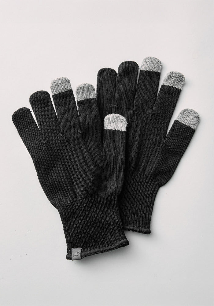 A pair of Asher Tech Gloves - Black by Woolx, featuring gray fingertips for touchscreen compatibility, is displayed on a light gray background. The gloves have ribbed cuffs and include a small tag on one glove.