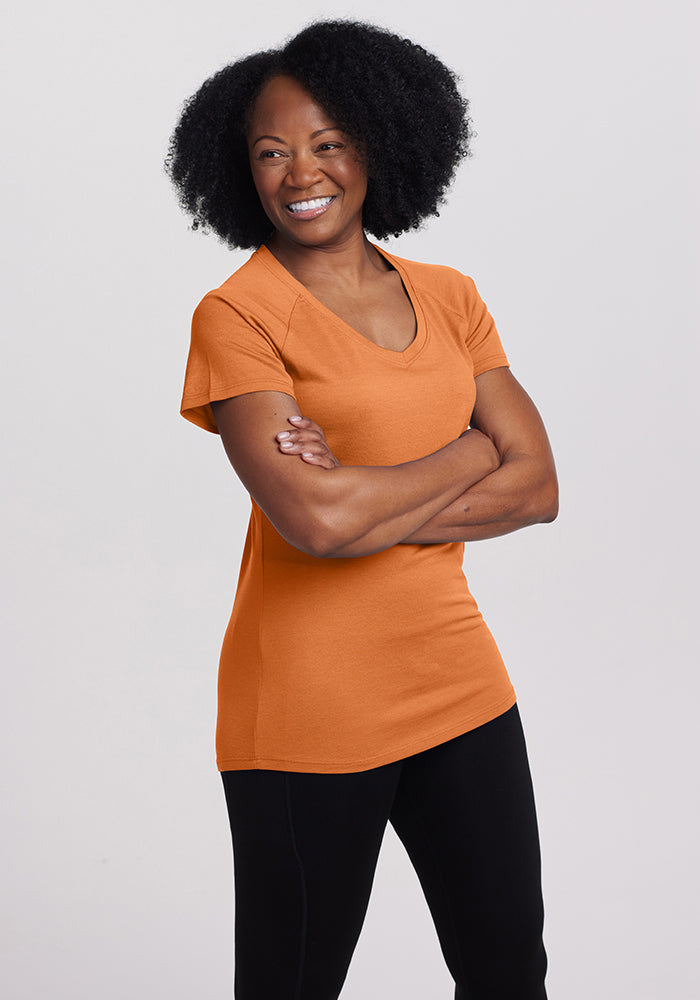 A person with curly hair smiles while standing with their arms crossed in a Woolx Mia Short Sleeve V Neck from the Stocking Stuffer Final Sale Deals! They are wearing orange and black pants against a plain background.