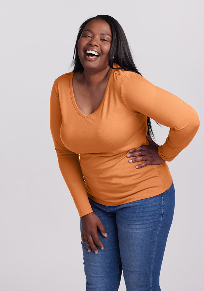 A person with long hair is delightedly smiling and laughing while wearing the Layla V Neck Top in orange from Woolx and blue jeans, standing against a plain background.