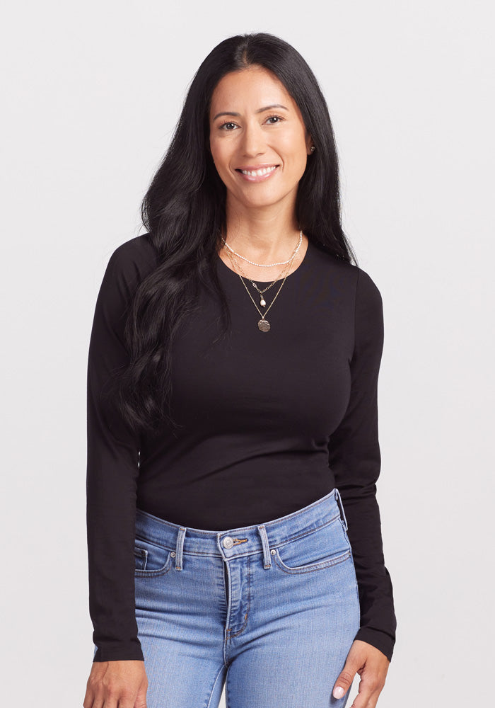 A woman with long black hair is smiling while wearing a chic black Sophia Bodysuit from Woolx and light blue jeans. She is also adorned with layered gold necklaces. The background is plain and white. 