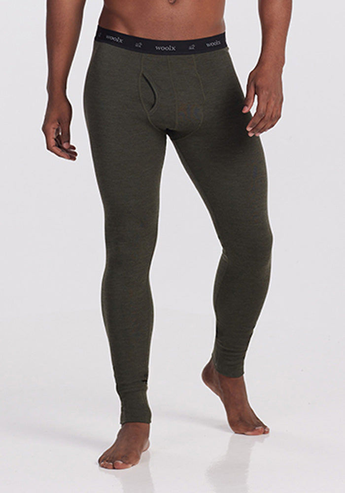 A person wearing the Woolx Backcountry Leggings in Dark Moss, designed for cold weather adventures. The waistband is black with the brand name "Woolx" repeated in gray. The individual, clad in this Merino wool base layer, stands against a plain background with hands relaxed by their sides. Only the lower torso to feet are visible. 