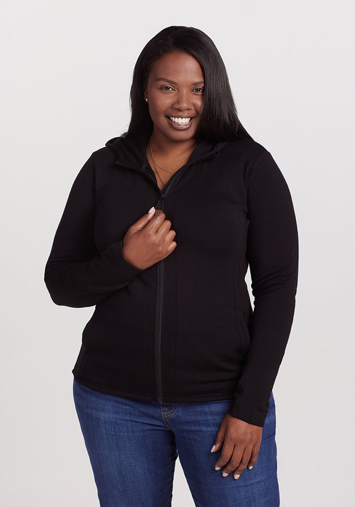 A person with long dark hair smiles while wearing the Woolx Zoey Zip Up Hoodie - Black and blue jeans. They are standing against a plain, light-colored background. 