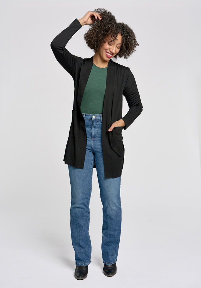 A person with curly hair smiles and poses against a plain background, wearing a green top, the Paisley Cardigan - Black by Woolx, blue jeans, and black shoes. One hand is in their pocket while the other playfully rests against their head.