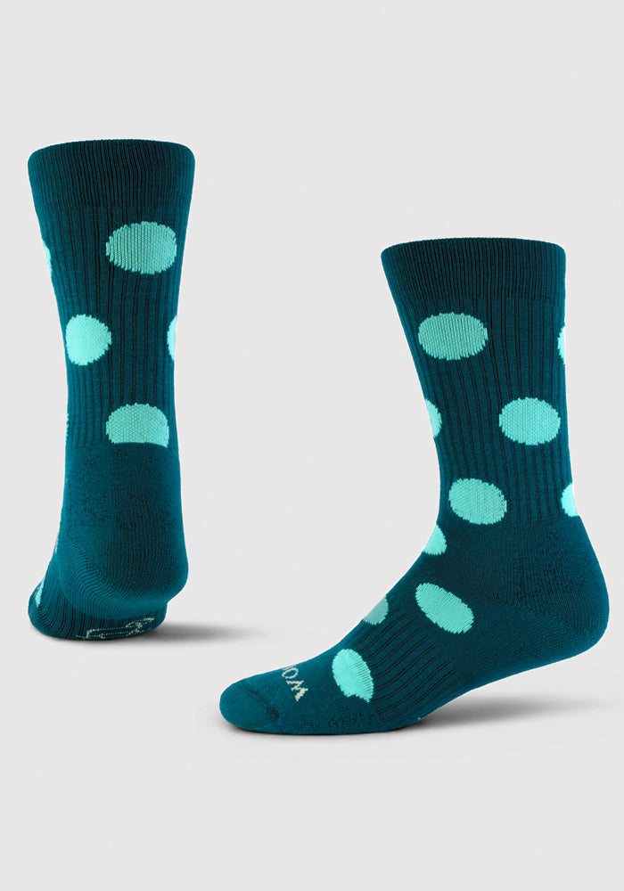 Product Shot: The Dotalicious Light Cushion Crew Socks by Woolx showcase two socks in teal, made from odor-resistant Merino wool, adorned with large light blue polka dots. One sock is positioned upright, while the other lies flat against a plain light gray background.