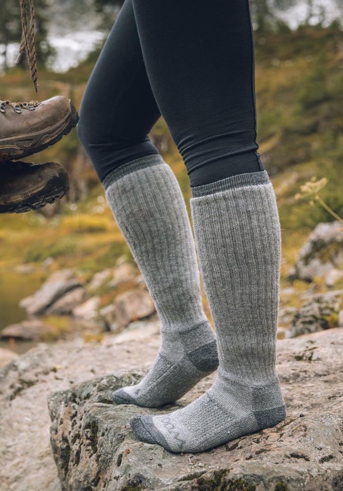 Dressed in Woolx Extreme Full Cushion Over The Calf socks in Charcoal Heather and black leggings, someone stands on a rocky terrain outdoors. Another individual's hiking boots can be seen nearby. The blurred green background suggests a natural winter haven ideal for seasonal adventures.