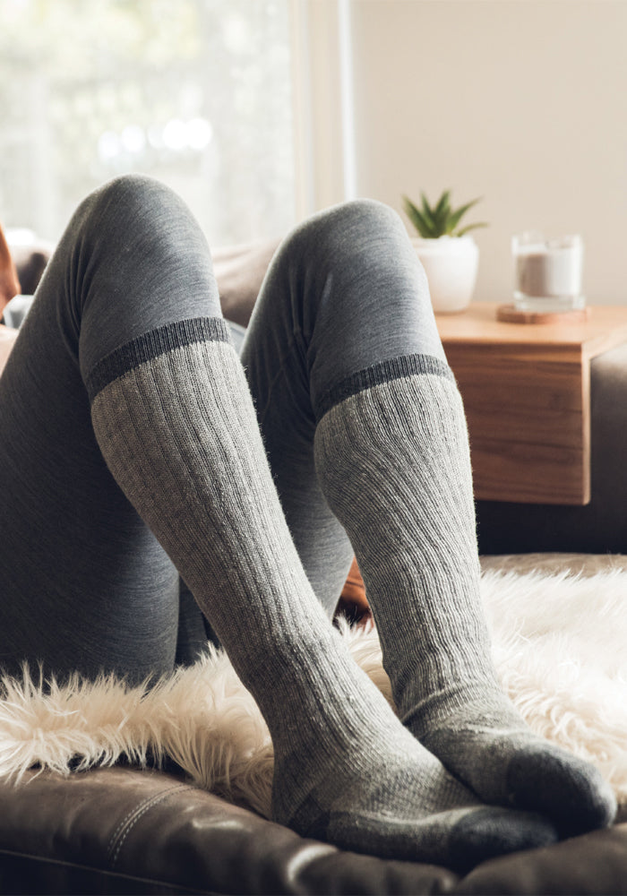 A person wearing Woolx's Extreme Full Cushion Over The Calf socks in Charcoal Heather is relaxing on a soft, white fur rug. The setting includes a wooden side table with a plant and candle, complemented by the gentle natural light streaming through the window—perfect for cozy winter activities at home.