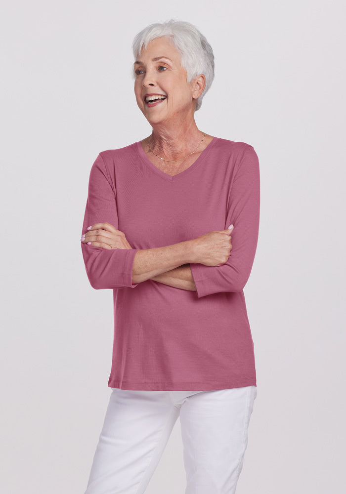 An older woman with short white hair is smiling with her arms crossed, dressed in a pink Elena V Neck - Mesa Rose top from Woolx and white pants. The lightweight 150g/m² fabric ensures comfort, while the plain white background accentuates her serene confidence.