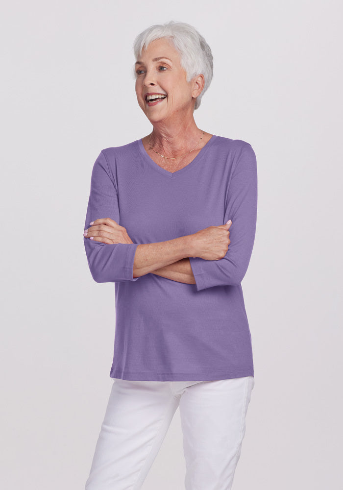 An older woman with short white hair is smiling and standing with her arms crossed. She is wearing a Montana Grape Elena V Neck top by Woolx, made from lightweight fabric, along with white pants, set against a plain gray background.