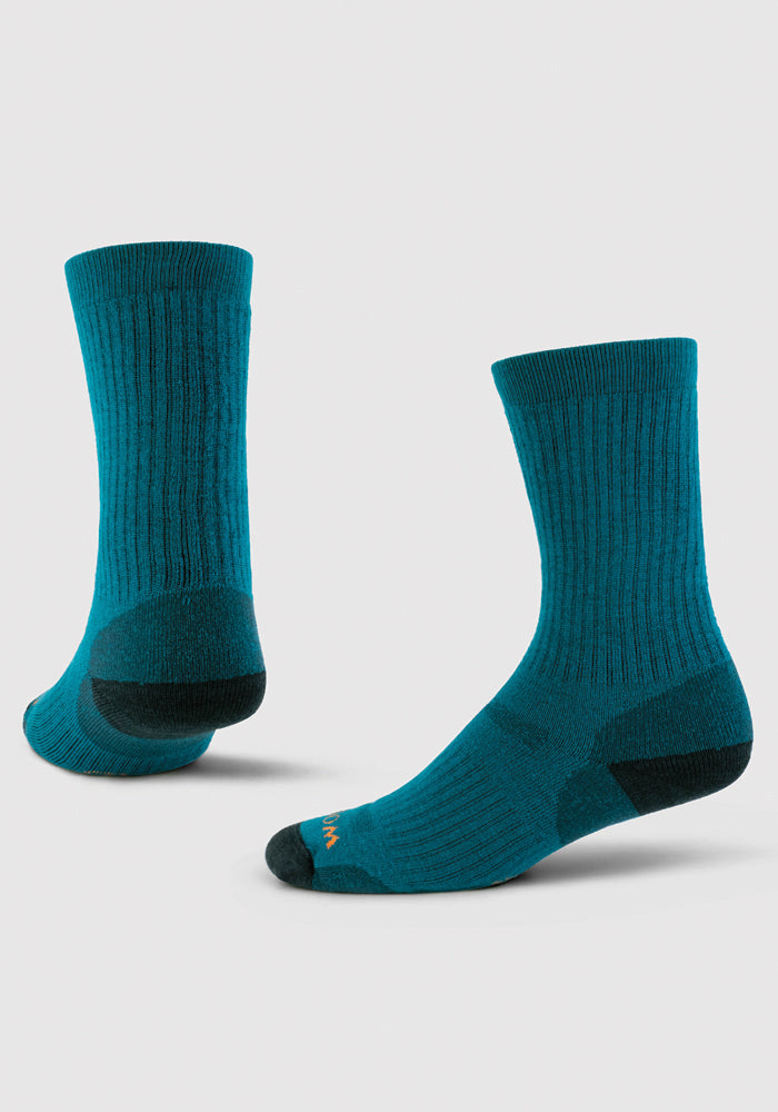 The Woolx Core Crew Sock Full Cushion in Real Teal features two socks made from moisture-wicking Merino wool with dark green reinforced heels and toes. The socks are displayed upright against a plain white background, highlighting their design and texture.