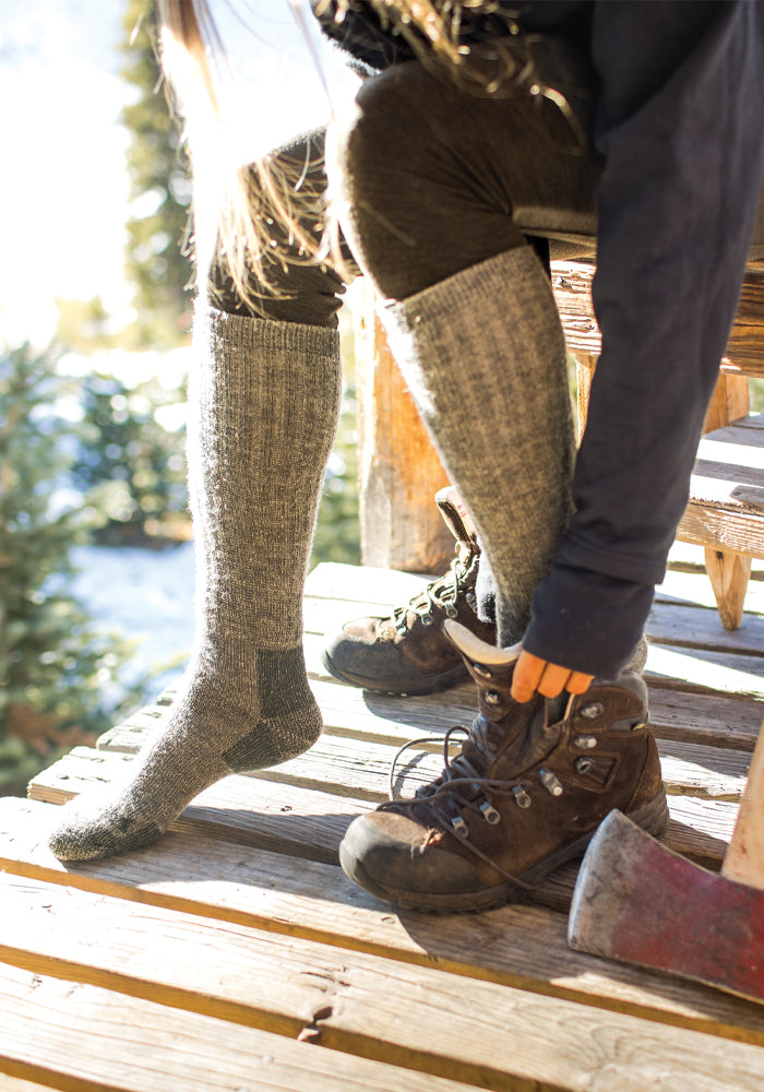 A person sits on wooden steps wearing Woolx's Extreme Full Cushion Over The Calf socks in Forest Dark Moss, while putting on brown hiking boots. Sunlight filters through the nearby trees, illuminating an axe resting on the steps.