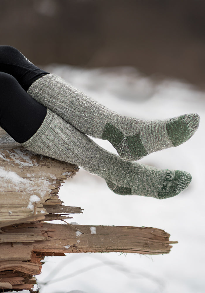 A person wearing the Woolx Extreme Full Cushion Over The Calf socks in Forest Dark Moss sits on a stack of firewood. Snow blankets the ground, setting a wintry outdoor scene. Their legs are crossed and clad in dark pants, perfect for experiencing moisture-wicking warmth in the chilly air.