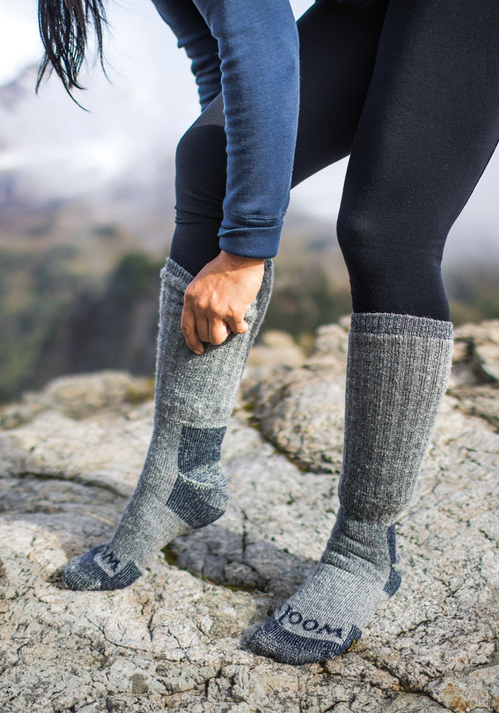 A person in a blue long-sleeve top and black leggings adjusts their Extreme Full Cushion Over The Calf - Navy Carbon socks from Woolx while standing on a rocky surface. The background is blurred, hinting at a mountainous or outdoor setting.