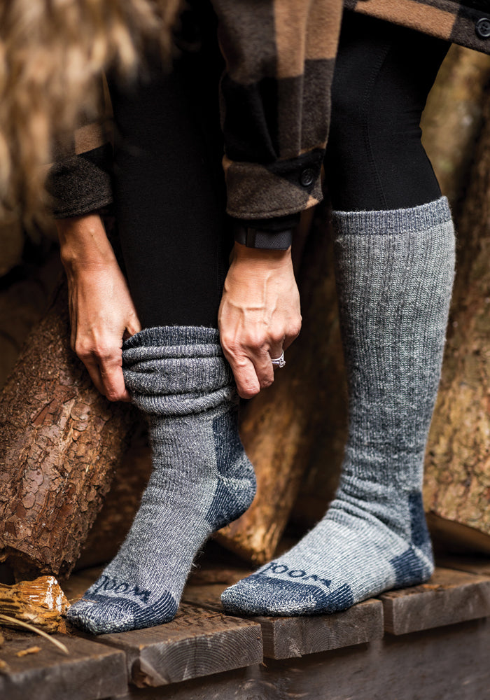 A person wearing a brown plaid coat adjusts a thick navy carbon merino wool sock from Woolx over black leggings. Sitting on stacked logs, with an over-the-calf design, one sock is rolled down while the other reaches up to the knee, offering heavyweight insulation against the chill.