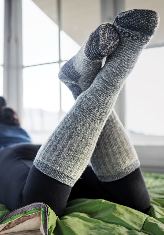 A person wearing navy carbon Extreme Full Cushion Over The Calf socks from Woolx is lying down with their legs crossed, comfortably relaxed on a green blanket. Sunlight filters through large windows in the background, suggesting a cozy indoor setting.