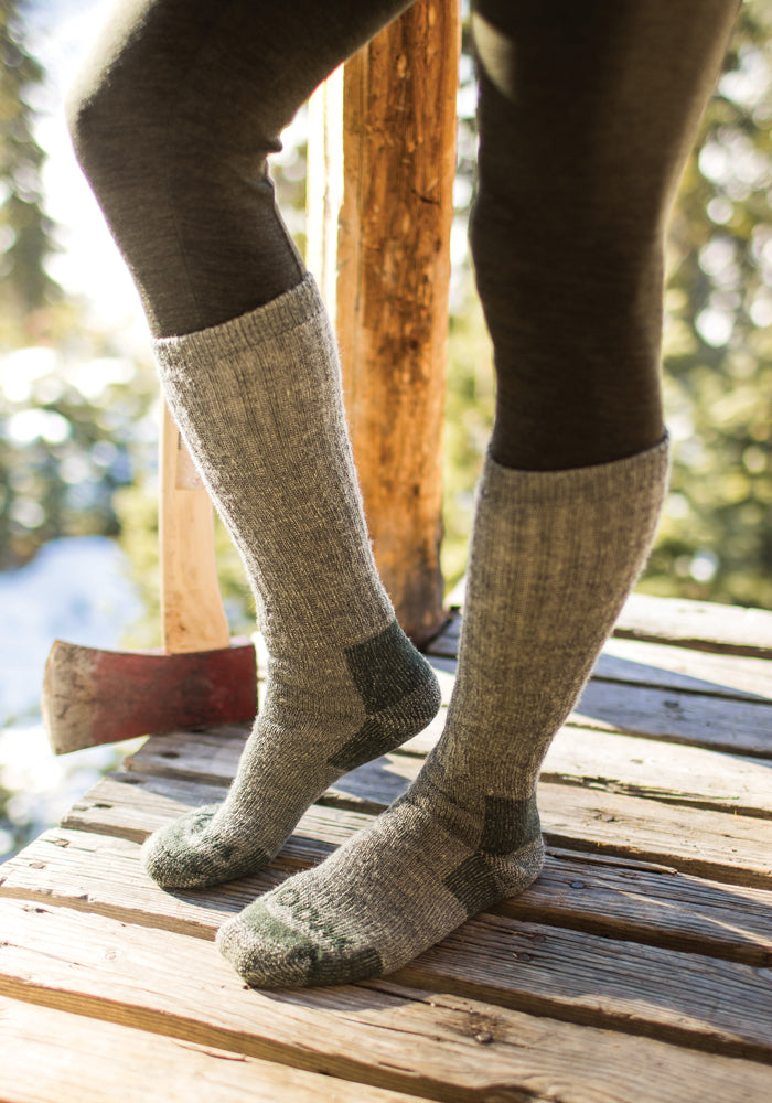 A person wearing Woolx's Extreme Full Cushion Over The Calf socks in Forest Dark Moss stands on a wooden porch. An axe is leaning against a nearby post. The scene unfolds outdoors with sunlight filtering through the trees, emphasizing the moisture-wicking comfort and over-the-calf design of their attire.