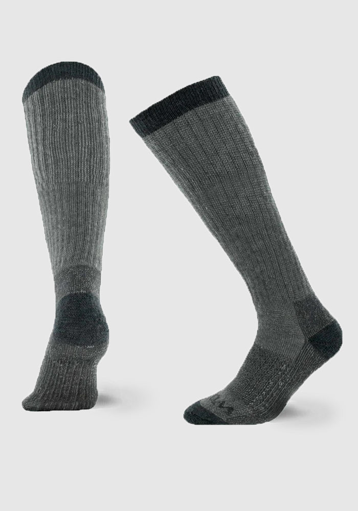 The product features the Extreme Full Cushion Over The Calf socks in Charcoal Heather from Woolx, depicted against a light background. One sock stands upright while the other is angled, highlighting its ribbed texture and cold-weather insulation.