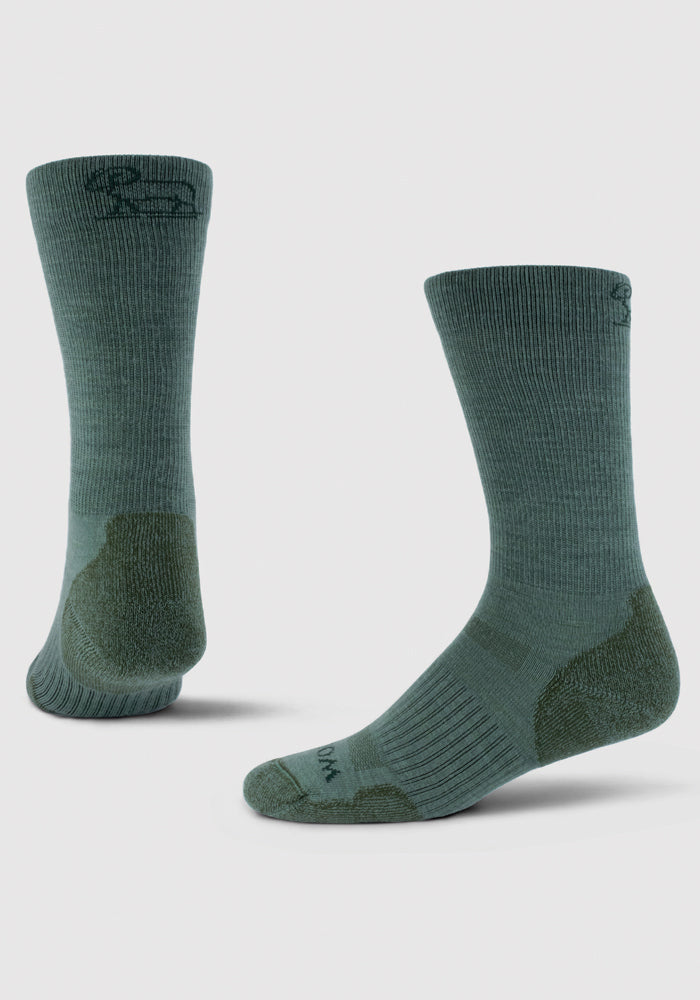 Presenting the Duck Green Harbor Crew Sock Light Cushion from Woolx, these socks feature a ribbed texture and a discreet logo on the top edge. Crafted from Merino wool, they offer moisture-wicking and odor-resistant properties. The mid-calf socks come with reinforced heels and toes for enhanced durability, displayed in side and back views against a plain white background.