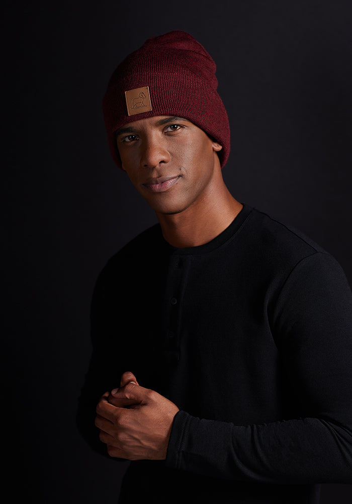 A person wearing a Woolx Baylor Beanie - Cranberry Melange and a black long-sleeve shirt stands against a dark background. They are looking directly at the camera with a neutral expression, hands clasped together.