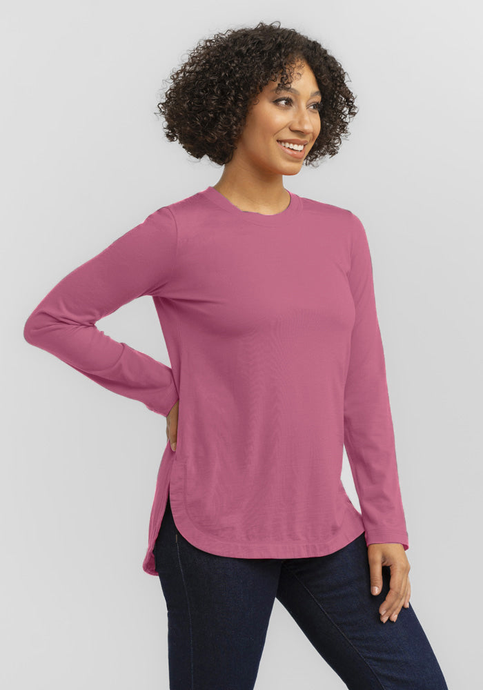 A curly-haired person smiles confidently with one hand on their hip, wearing a relaxed-fit Woolx Hazel Tunic in Mesa Rose and dark jeans against a plain white background. Note: Final Sale Deals!.