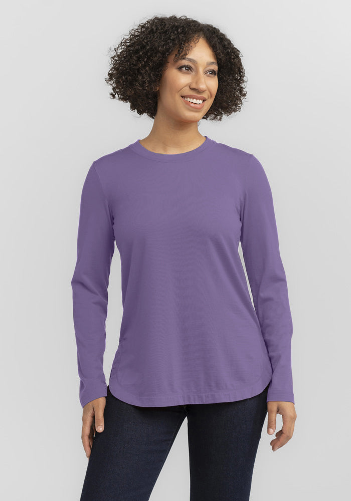 A person with curly hair smiles in a relaxed fit Hazel Tunic from Woolx, color Montana Grape, and dark jeans, standing against a plain white background.