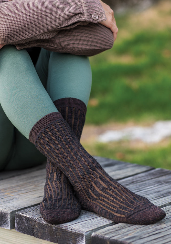 A person sits on a wooden bench, wearing a brown sweater, green leggings, and Woolx's Joey Full Cushion Mid Calf Socks in Dark Espresso. The person’s arms are folded over their knees as they enjoy the day. Green grass is visible in the background.