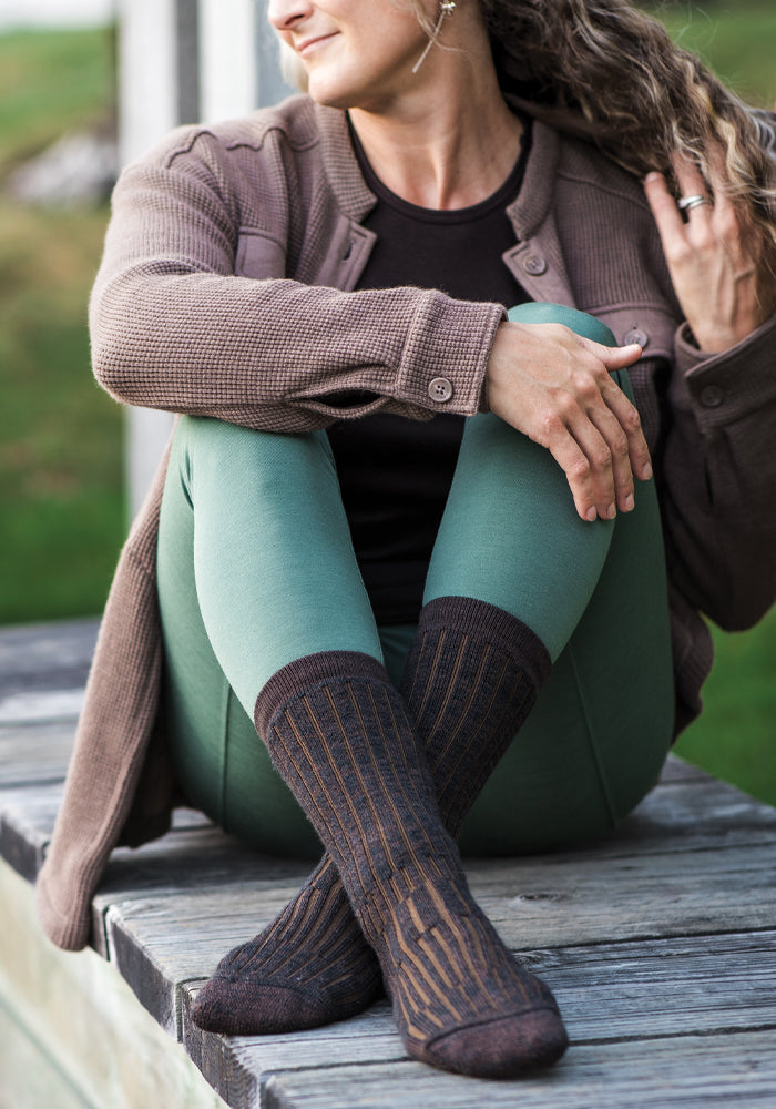 A person with curly hair sits outdoors on a wooden surface, wearing a brown jacket, black shirt, green leggings, and the warmest Joey Full Cushion Mid Calf Socks in Dark Espresso from Woolx. Their arms rest on their knees as they gaze into the distance with a relaxed expression.