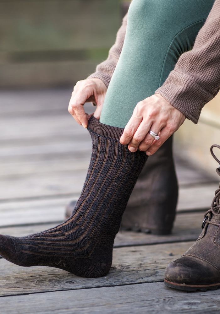 A person wearing green leggings and a brown sweater adjusts their Joey Full Cushion Mid Calf Socks in Dark Espresso from Woolx while sitting on a wooden surface. They are wearing brown lace-up boots and a ring on their left hand.