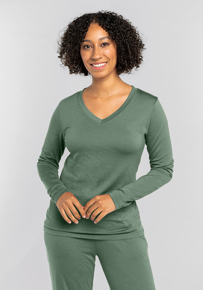 A person wearing Woolx's Lily V Neck Lounge Top in Duck Green, made from moisture-wicking Merino wool, smiles while standing against a gray background.