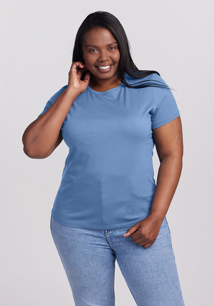 A smiling person with long hair is wearing the Woolx Liza Crew Neck in Coronet Blue and blue jeans, standing against a plain background. They have one hand on their hip and the other touching their neck.