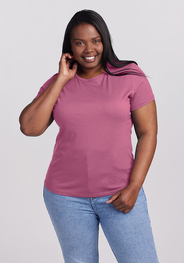 A woman with long dark hair is confidently smiling in a Mesa Rose Liza Crew Neck by Woolx, paired with light blue jeans. She gently touches her hair against a plain gray background that highlights her vibrant style.