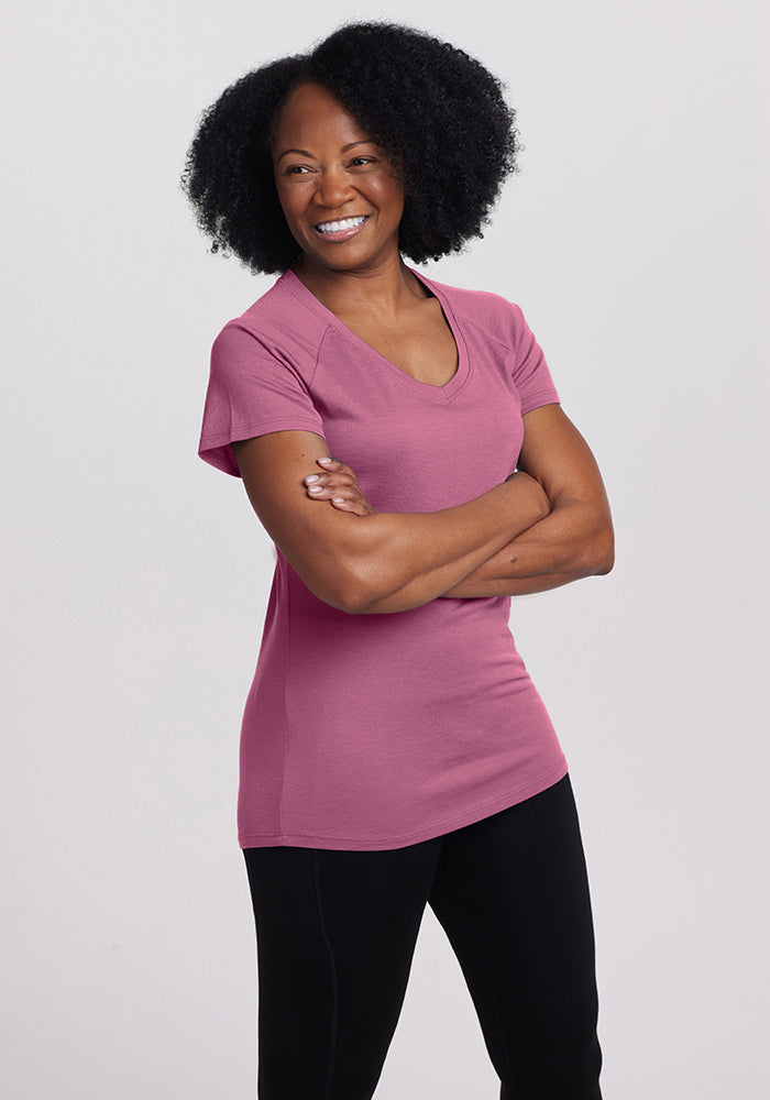 A person with curly black hair smiles while standing with their arms crossed, wearing a pink Mia Short Sleeve V Neck from Woolx and black pants against a plain light gray background.