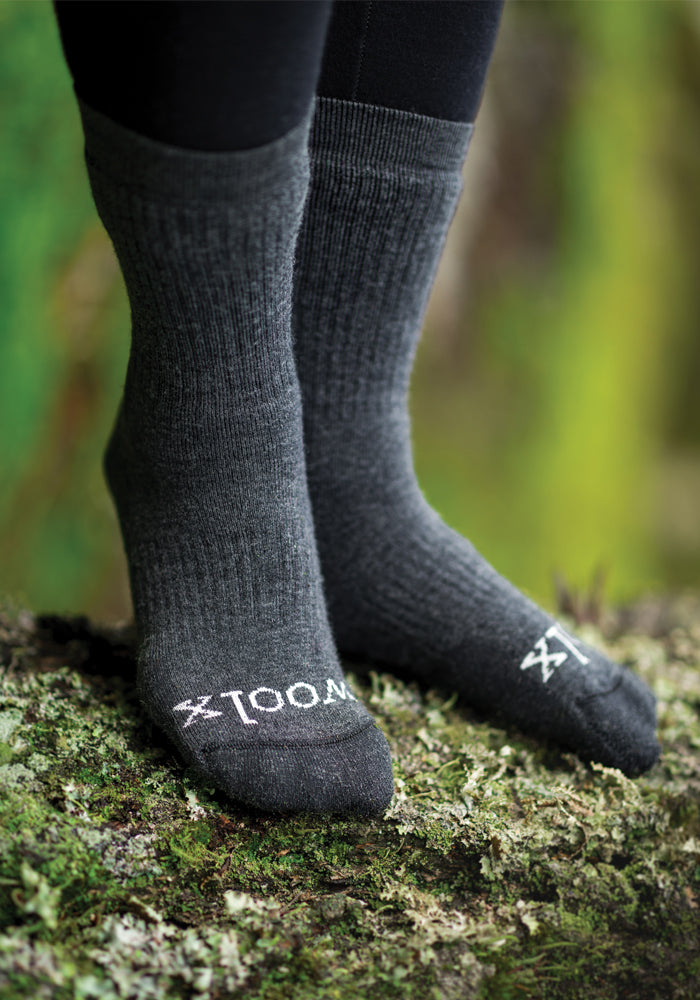 A close-up shot captures someone wearing the Milo Full Cushion Cold Weather Socks in Dark Grey Charcoal by Woolx, standing on a mossy surface. These socks, crafted from moisture-wicking Merino wool, feature a knit texture with "WOOL" displayed in white letters. The blurred greenery suggests an outdoor setting.