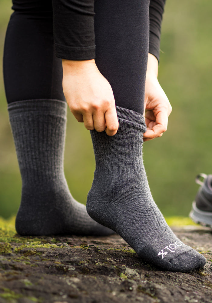 A person wearing Milo Full Cushion Cold Weather Socks in dark grey charcoal by Woolx stands outdoors on a moss-covered rock. Their hands adjust the moisture-wicking socks, against a lush and green blurred background.