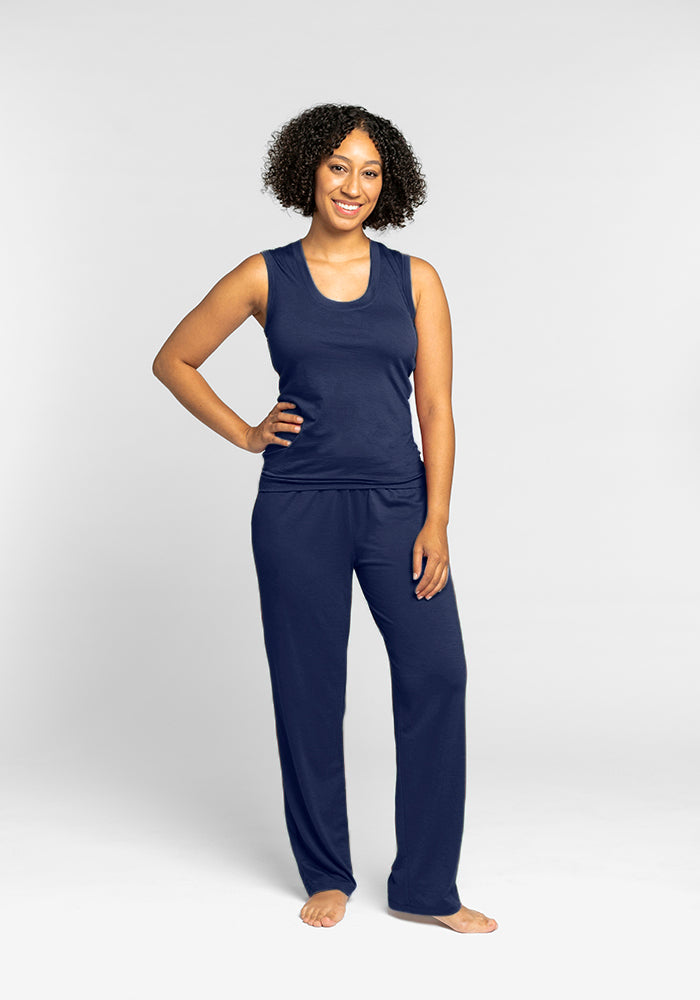 A person with curly hair smiles while standing against a neutral background. They are wearing the Phoebe Lounge Tank in Starry Night from Woolx and matching pants crafted from temperature-regulating Merino wool, barefoot with one hand on their hip.