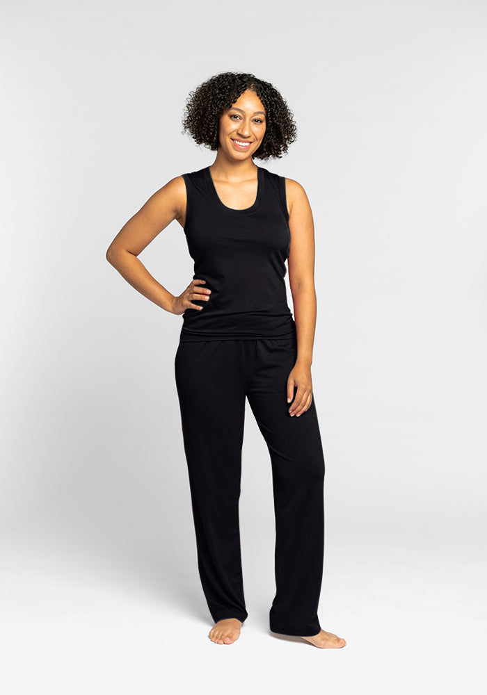 A person with curly hair is smiling and posing with one hand on their hip, wearing a sleek, sleeveless black Woolx Phoebe Lounge Tank and matching black pants. Standing barefoot against a plain white background, they perfectly capture the essence of travel-friendly clothing.