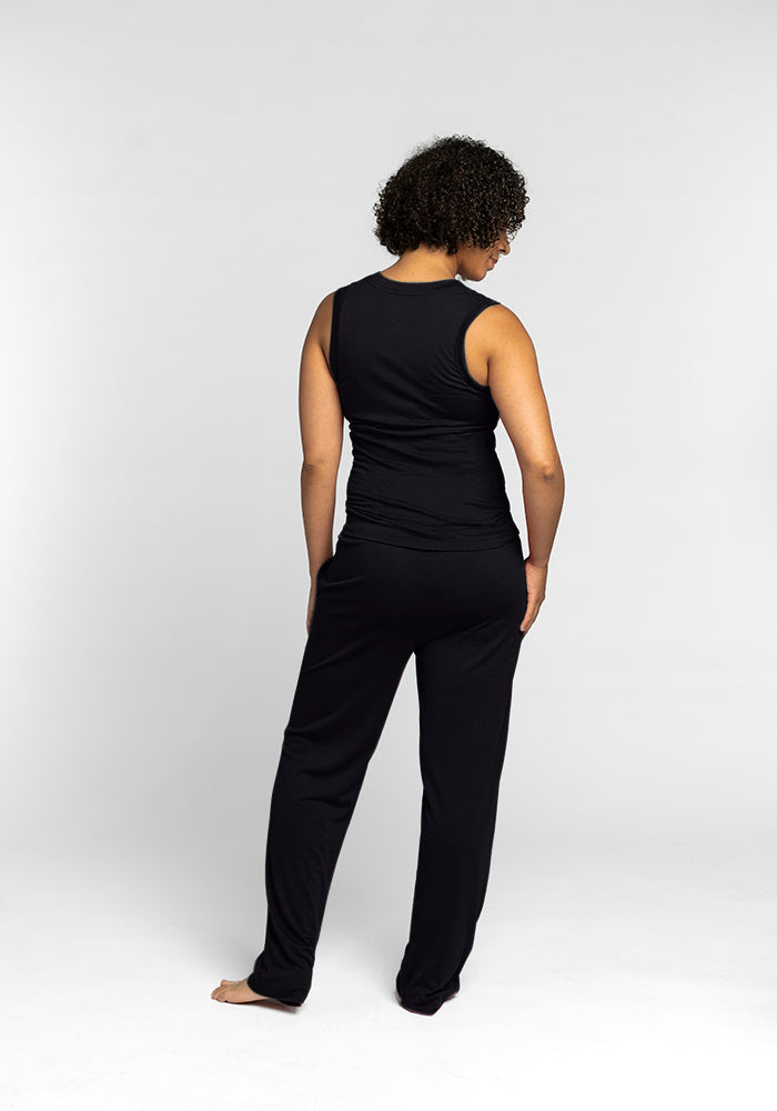 A person with curly hair, dressed in a travel-friendly Phoebe Lounge Tank - Black from Woolx and matching black pants, stands barefoot on a light gray background. Facing away from the camera with their head slightly turned to the side, they exude effortless style while enjoying the comfort of Australian Merino wool.