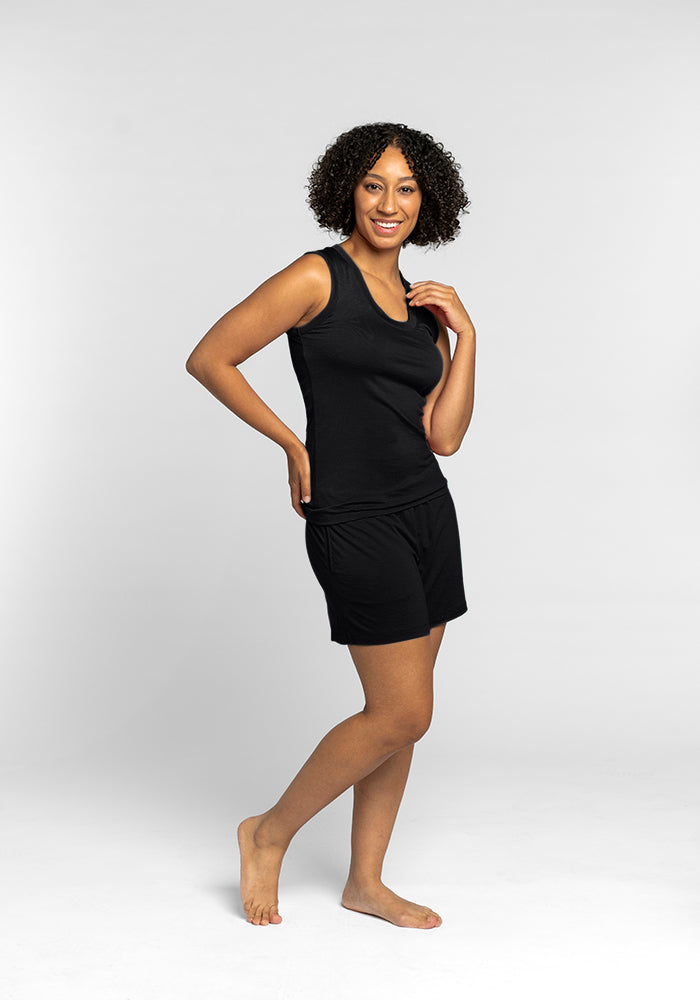 A person with curly hair is smiling, barefoot and posed against a plain white background, wearing the travel-friendly Phoebe Lounge Tank in black by Woolx along with shorts.